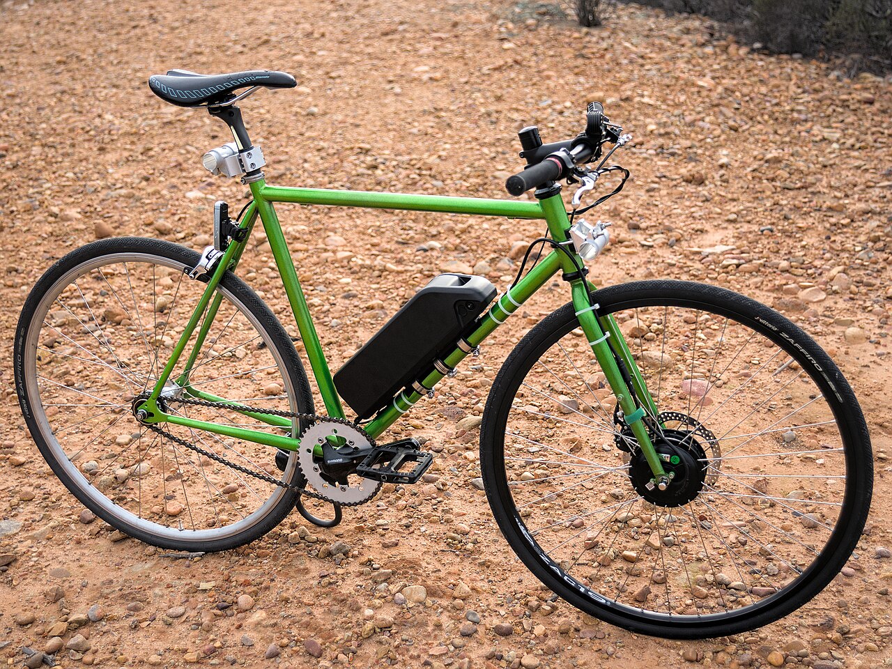Green road bicycle on gravel, fitted with a rear hub motor and a downtube battery.